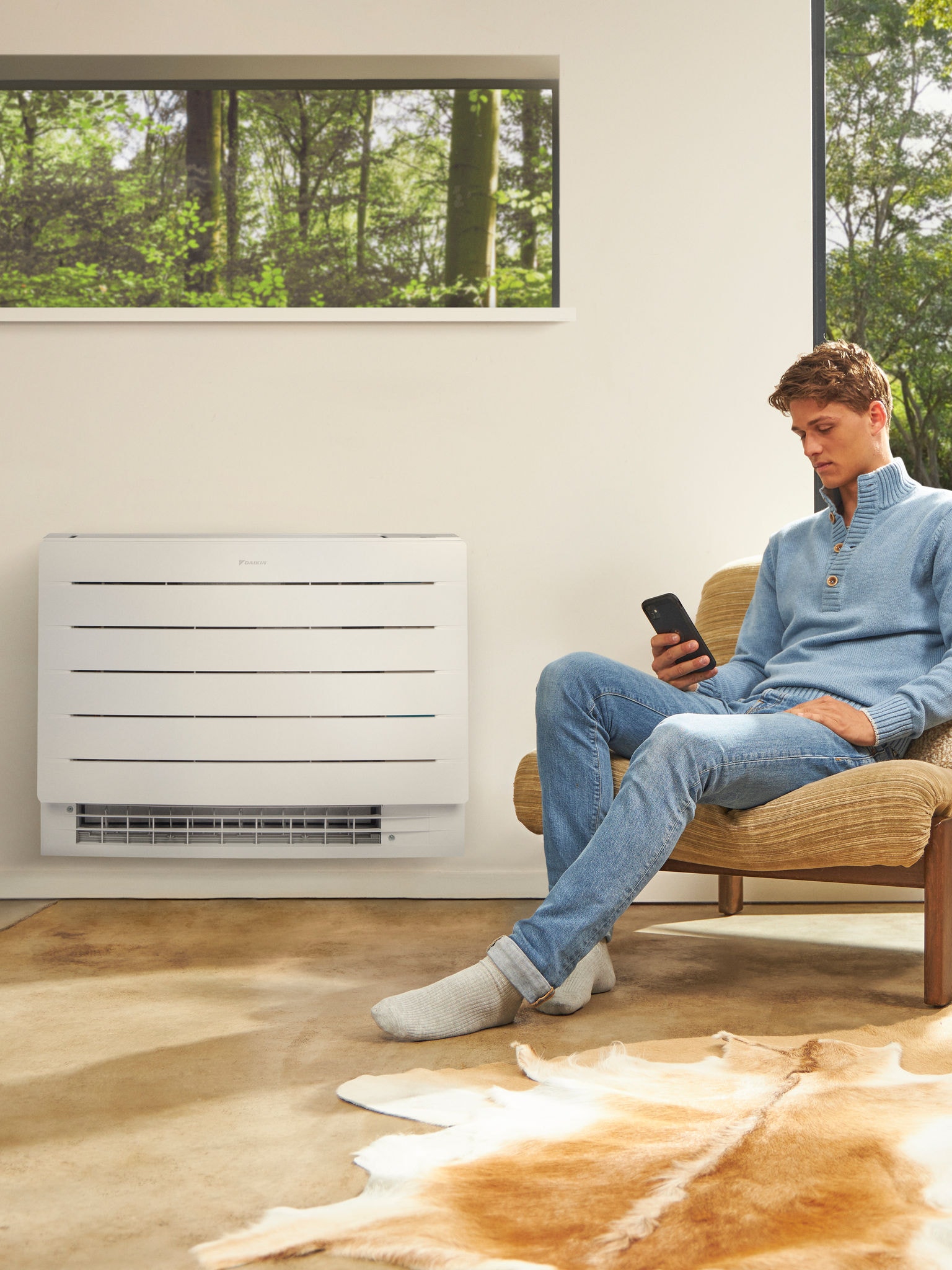 Homem sentado numa sala luminosa a olhar para o telefone com uma bomba de calor na parede atrás dele