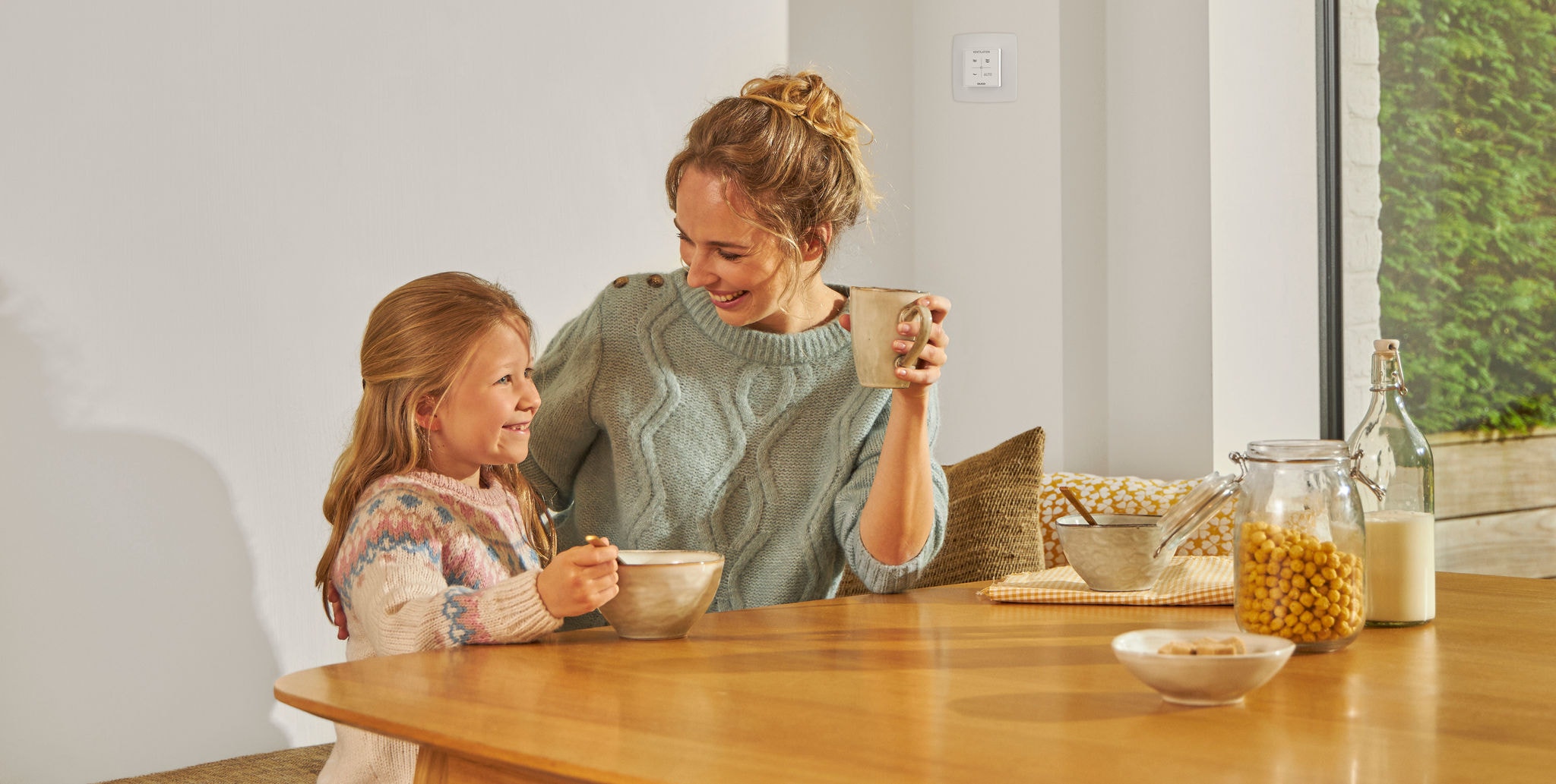 Uma menina a comer de uma tigela sentada à mesa ao lado de uma mulher com uma caneca na mão, ambas a sorrir, felizes e relaxadas, a desfrutar de ar saudável.