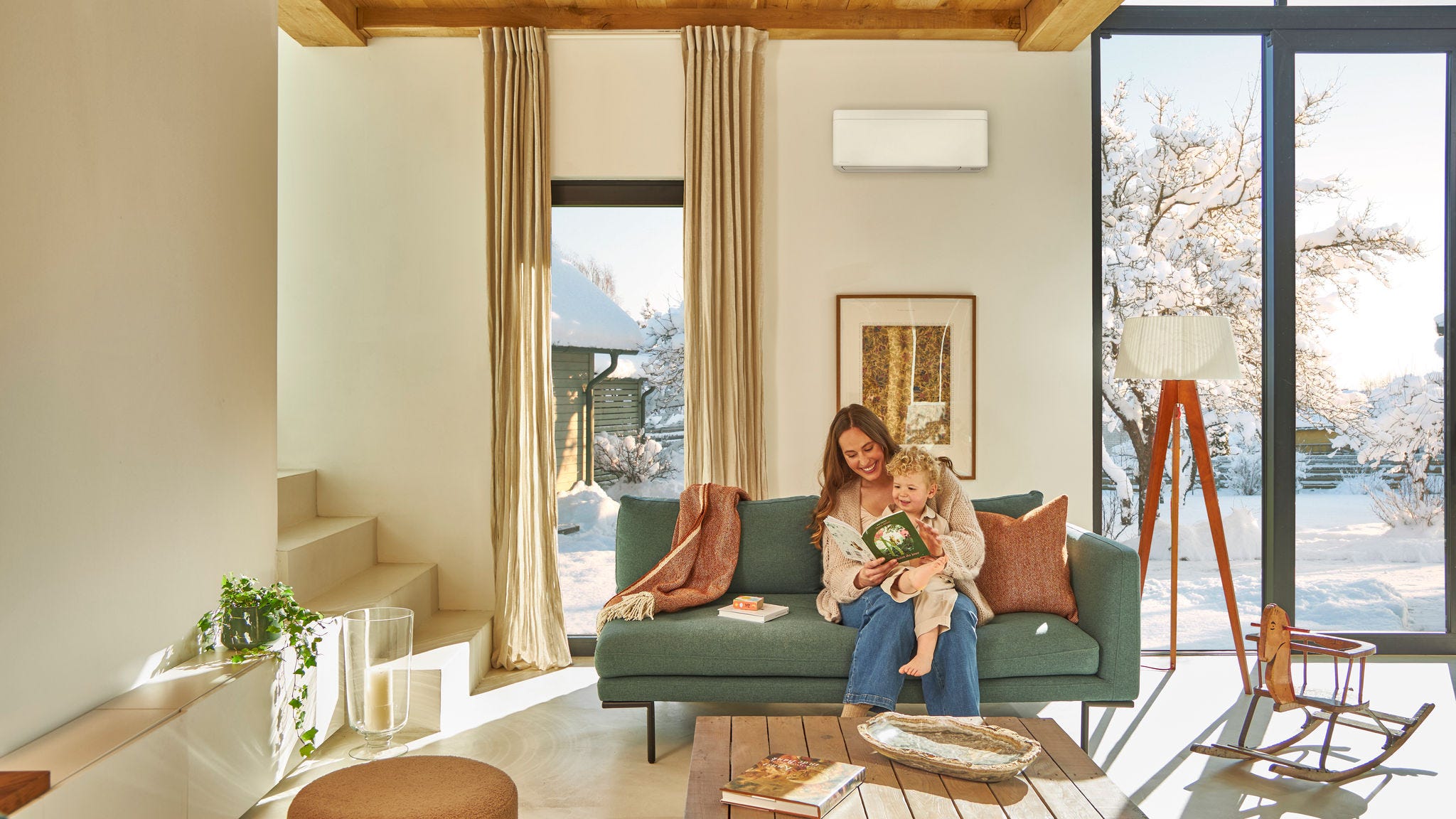 Mãe sentada no sofá sob uma bomba de calor ar-ar montada na parede lê um livro infantil com o filho no colo, desfrutando do calor enquanto neva lá fora